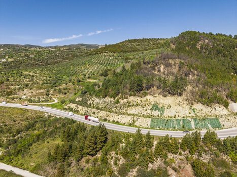 Aerial view of a winding road through the Vjosa Wild River National Park with a red truck and vehicles surrounded by green hills and terraced slopes Permet Gjirokaster County Albania. Aerial view of a winding road through the Vjosa Wild River National Park with a red truck and vehicles surrounded by green hills and terraced slopes Permet Gjirokaster County Albania.