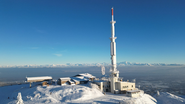 Aerial view of Col de la Faucille featuring a tall telecommunications tower and snow covered buildings on a mountain peak under a clear blue sky in Gex Auvergne Rhone Alpes France. Aerial view of Col de la Faucille featuring a tall telecommunications tower and snow covered buildings on a mountain peak under a clear blue sky in Gex Auvergne Rhone Alpes France.