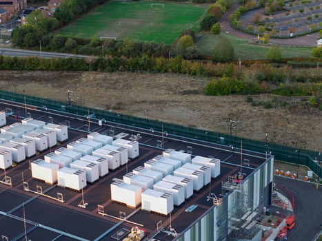 Aerial view of a data center with rows of white cooling units on a flat roof adjacent to a green fence grassy field and parking area in the United Kingdom. Aerial view of a data center with rows of white cooling units on a flat roof adjacent to a green fence grassy field and parking area in the United Kingdom.
