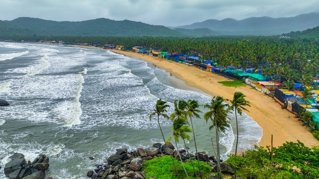 Aerial view of Palolem Beach with palm trees sandy shore colorful beach shacks waves and lush green hills under a cloudy sky in Canacona Goa India.