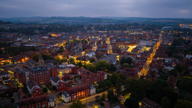 Aerial view of Salisbury at dusk featuring illuminated historic buildings winding streets and the iconic cathedral spire rising above the city rooftops in Salisbury England United Kingdom. Aerial view of Salisbury at dusk featuring illuminated historic buildings winding streets and the iconic cathedral spire rising above the city rooftops in Salisbury England United Kingdom.