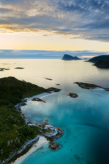 Aerial view of the turquoise waters and rocky coastline of Sommaroy with distant mountains under a soft sunset sky in Sommaroy Troms Norway. Aerial view of the turquoise waters and rocky coastline of Sommaroy with distant mountains under a soft sunset sky in Sommaroy Troms Norway.