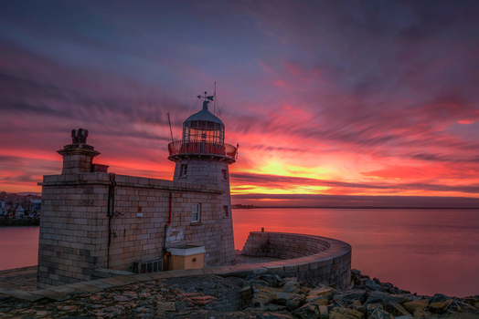 View of Howth Harbour lighthouse at sunset with vibrant pink and orange clouds over the calm sea in Ireland. View of Howth Harbour lighthouse at sunset with vibrant pink and orange clouds over the calm sea in Ireland.