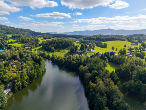 Aerial view of Fojtka Reservoir surrounded by lush green forests rolling hills and scattered houses under a bright blue sky with white clouds in Mnisek Liberecky kraj Czechia. Aerial view of Fojtka Reservoir surrounded by lush green forests rolling hills and scattered houses under a bright blue sky with white clouds in Mnisek Liberecky kraj Czechia.