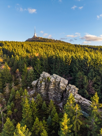 Aerial view of the Stone Gate rock formation surrounded by dense forest with the Jested Tower on the mountain peak under a clear blue sky in Svetla pod Jestedem Liberec Region Czechia. Aerial view of the Stone Gate rock formation surrounded by dense forest with the Jested Tower on the mountain peak under a clear blue sky in Svetla pod Jestedem Liberec Region Czechia.