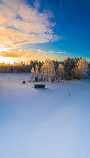 Aerial view of a small wooden structure in a vast snow covered field surrounded by a forest of frost covered trees under a vibrant sunset sky in Latvia. Aerial view of a small wooden structure in a vast snow covered field surrounded by a forest of frost covered trees under a vibrant sunset sky in Latvia.