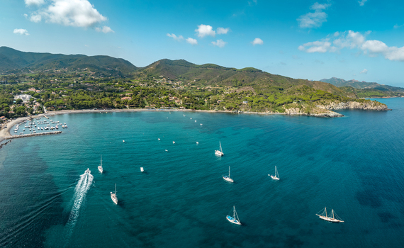 Aerial view of Lacona Beach with sailboats and motorboats anchored in the turquoise bay surrounded by lush green hills and coastal vegetation in Lacona Tuscany Italy.