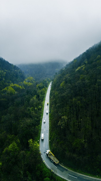 Aerial view of a winding road cuts through a dense forest with vehicles dotting the landscape under a misty sky Novi Sad Vojvodina Serbia. Aerial view of a winding road cuts through a dense forest with vehicles dotting the landscape under a misty sky Novi Sad Vojvodina Serbia.