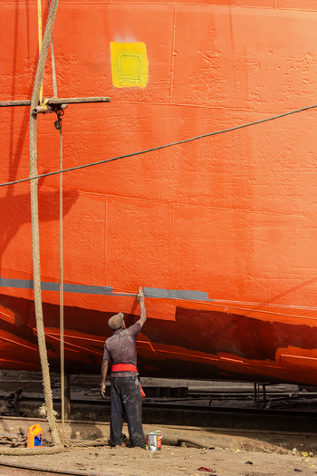 Keraniganj Bangladesh 12 February 2024 View of a worker painting the massive orange hull of a ship at the shipyard with ropes and paint cans visible.