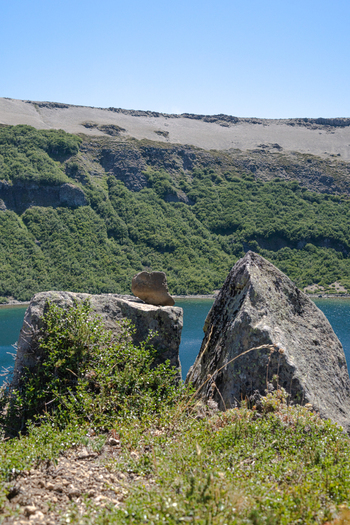 View of Volcan Batea Mahuida with large volcanic rocks in the foreground and a serene lake nestled against the green slopes of the volcano in Alumine Neuquen Argentina. View of Volcan Batea Mahuida with large volcanic rocks in the foreground and a serene lake nestled against the green slopes of the volcano in Alumine Neuquen Argentina.