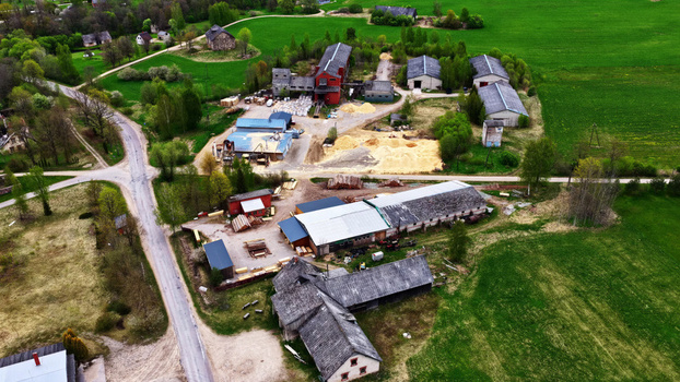 Aerial view of a rural sawmill facility featuring industrial buildings piles of wood chips and timber storage surrounded by green fields trees and a country road Latvija. Aerial view of a rural sawmill facility featuring industrial buildings piles of wood chips and timber storage surrounded by green fields trees and a country road Latvija.