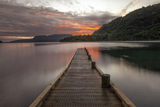 View of Lake Tarawera with a wooden jetty extending into the calm water at sunset featuring soft clouds and distant hills in Rotorua Bay of Plenty Region New Zealand.