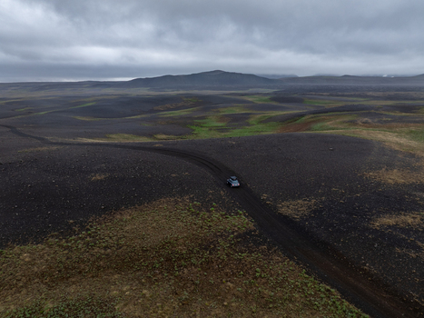 Aerial view of a car driving along a dark volcanic dirt road through a vast barren landscape with patches of green moss and distant mountains under a cloudy sky in Iceland. Aerial view of a car driving along a dark volcanic dirt road through a vast barren landscape with patches of green moss and distant mountains under a cloudy sky in Iceland.