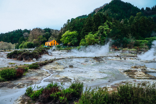 View of steaming vents rising from the ground amidst lush greenery with a bright yellow house perched on a hillside in Furnas Azores Portugal.
