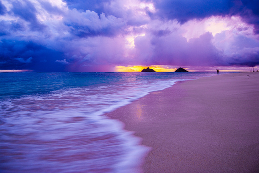 View of Lanikai Beach at sunset with dramatic purple and orange clouds gentle waves washing onto the sandy shore and the Mokulua Islands on the horizon Kailua Hawaii United States.