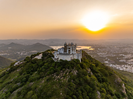 Aerial view of Sajjangarh Palace on a hilltop at sunset with lush slopes and a shimmering lake in the distance Udaipur Rajasthan India.