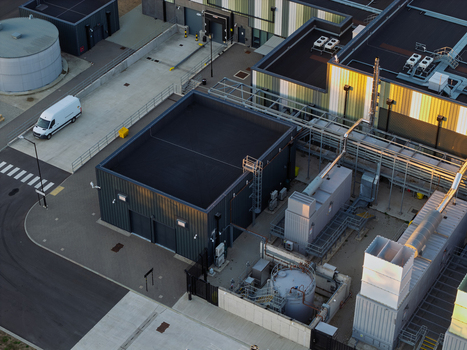 Aerial view of a modern data center facility featuring industrial cooling units technical infrastructure and a white van parked on the paved grounds in the United Kingdom. Aerial view of a modern data center facility featuring industrial cooling units technical infrastructure and a white van parked on the paved grounds in the United Kingdom.
