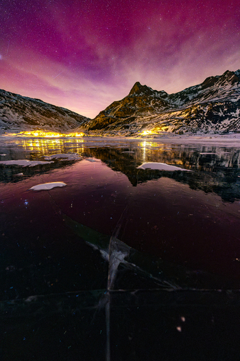 View of a frozen lake reflecting a vibrant pink and purple night sky with snowy mountains and distant city lights in Madesimo Lombardy Italy. View of a frozen lake reflecting a vibrant pink and purple night sky with snowy mountains and distant city lights in Madesimo Lombardy Italy.