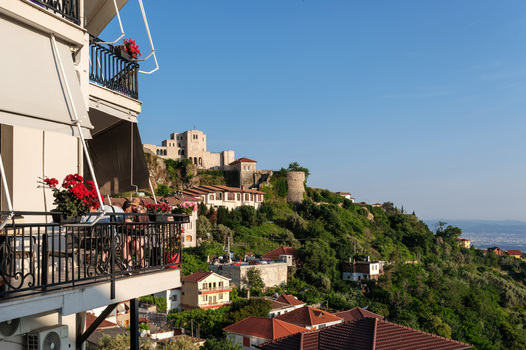 View of Kruja Castle on a lush green hill with traditional houses and people on a balcony with red flowers in the foreground Kruje Durres County Albania.