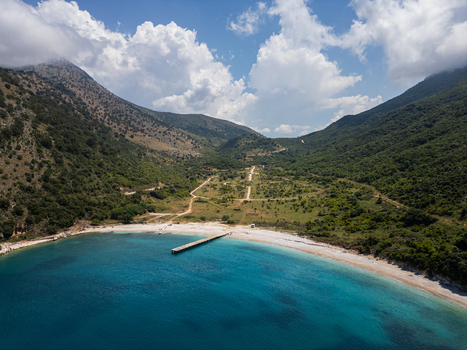 Aerial view of Kakome Bay featuring turquoise waters a wooden pier and a white pebble beach nestled between lush green mountains in Vlore Vlore County Albania. Aerial view of Kakome Bay featuring turquoise waters a wooden pier and a white pebble beach nestled between lush green mountains in Vlore Vlore County Albania.