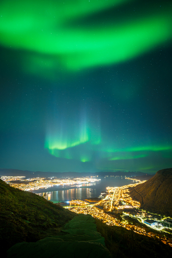 View of the vibrant green aurora borealis illuminating the night sky above the city lights of Tromso Troms Norway. View of the vibrant green aurora borealis illuminating the night sky above the city lights of Tromso Troms Norway.
