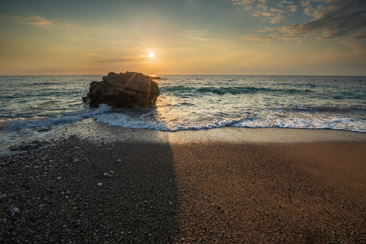 View of sun kissing the horizon and a large rock while waves gently wash over the sandy shore Kathisma beach Kathisma Lefkada Greece. View of sun kissing the horizon and a large rock while waves gently wash over the sandy shore Kathisma beach Kathisma Lefkada Greece.