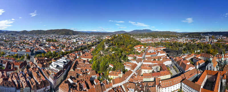 Aerial view of sun kissed red rooftops nestled amidst lush greenery framed by distant mountains and a clear blue sky Graz Styria Austria.
