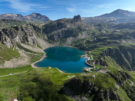 Aerial view of Agnel Lake surrounded by rugged mountain peaks and winding alpine roads under a clear blue sky Ceresole Reale Piedmont Italy.