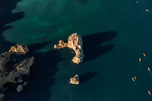 Aerial view of limestone rock formations rising from the turquoise Atlantic Ocean with colorful kayaks paddling nearby in Lagos Faro District Portugal.