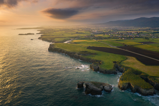 Aerial view of the rugged coastline of Gaztelugatxe featuring steep cliffs lush green fields and the Atlantic Ocean under a warm sunset sky Gaztelugatxeko Doniene Basque Country Spain. Aerial view of the rugged coastline of Gaztelugatxe featuring steep cliffs lush green fields and the Atlantic Ocean under a warm sunset sky Gaztelugatxeko Doniene Basque Country Spain.