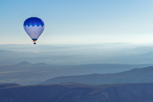 Aerial view of a blue and white hot air balloon floating over the misty volcanic landscape and layered ridges of Cappadocia in Kayseri Kayseri Turkiye. Aerial view of a blue and white hot air balloon floating over the misty volcanic landscape and layered ridges of Cappadocia in Kayseri Kayseri Turkiye.