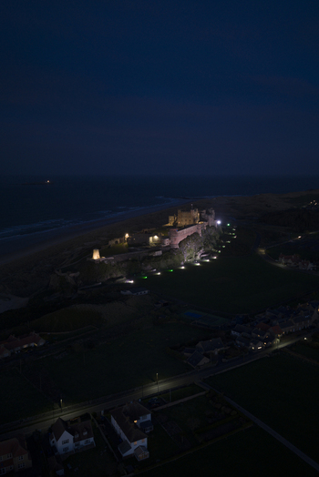 Aerial view of Bamburgh Castle illuminated at night overlooking the North Sea coast and village rooftops Bamburgh England United Kingdom. Aerial view of Bamburgh Castle illuminated at night overlooking the North Sea coast and village rooftops Bamburgh England United Kingdom.