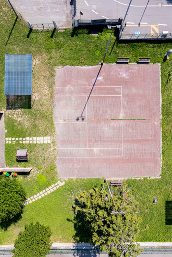 Aerial view of a worn reddish brown tennis court surrounded by lush green grass and trees in Turin Piedmont Italy. Aerial view of a worn reddish brown tennis court surrounded by lush green grass and trees in Turin Piedmont Italy.
