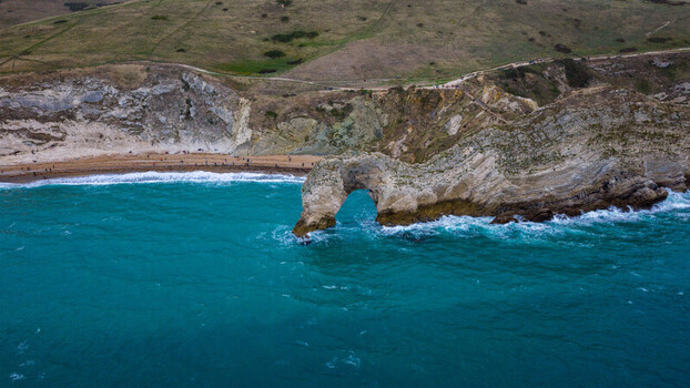 Aerial view of Durdle Door a natural limestone arch on the Jurassic Coast with turquoise sea waves crashing against cliffs and people on the beach in Wareham England United Kingdom. Aerial view of Durdle Door a natural limestone arch on the Jurassic Coast with turquoise sea waves crashing against cliffs and people on the beach in Wareham England United Kingdom.