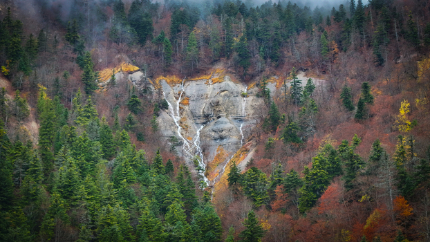 Aerial view of a cascade cutting through rugged rock face amidst a tapestry of evergreens and autumnal hues along Route du Creux du Cerf Gex Auvergne Rhône Alpes France. Aerial view of a cascade cutting through rugged rock face amidst a tapestry of evergreens and autumnal hues along Route du Creux du Cerf Gex Auvergne Rhône Alpes France.