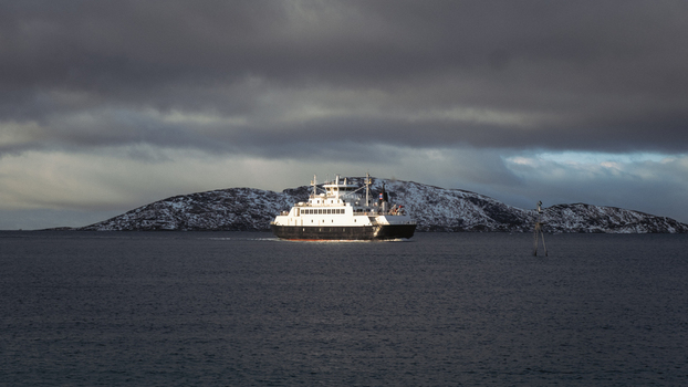 View of a ferry cutting through the still dark waters toward a snow dusted island under a moody overcast sky Brensholmen Troms Norway. View of a ferry cutting through the still dark waters toward a snow dusted island under a moody overcast sky Brensholmen Troms Norway.