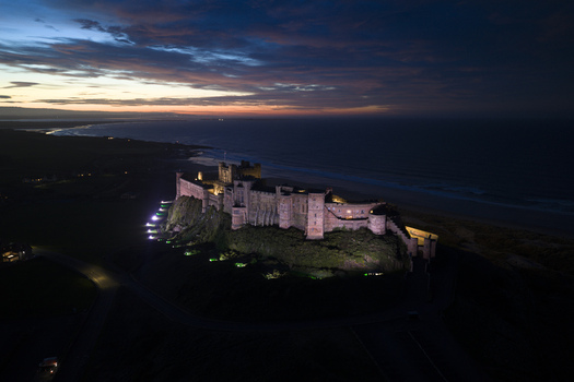 Aerial view of Bamburgh Castle illuminated at night on a rocky cliff overlooking the North Sea coastline under a twilight sky in Bamburgh England United Kingdom. Aerial view of Bamburgh Castle illuminated at night on a rocky cliff overlooking the North Sea coastline under a twilight sky in Bamburgh England United Kingdom.