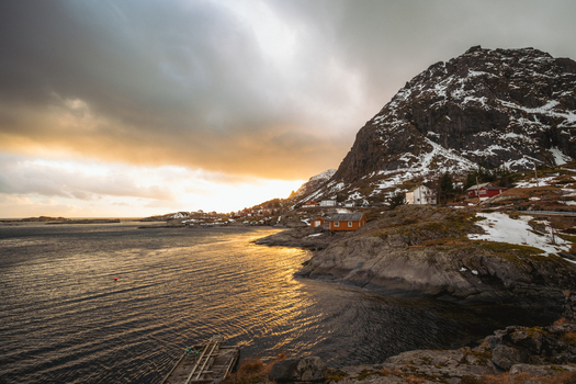 View of golden light kissing the tranquil waters near rocky shores contrasting with snow dusted peaks and quaint red roofed houses Tind Nordland Norway.