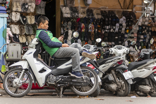 Hanoi Ha Noi Vietnam 11 November 2025 View of a man sitting on a white motorbike while checking his phone in front of a footwear shop.