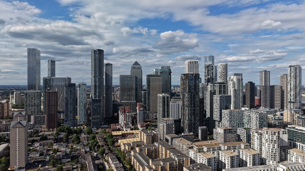 London United Kingdom 16 June 2026 Aerial view of the Canary Wharf financial district skyline with modern skyscrapers under a cloudy blue sky.