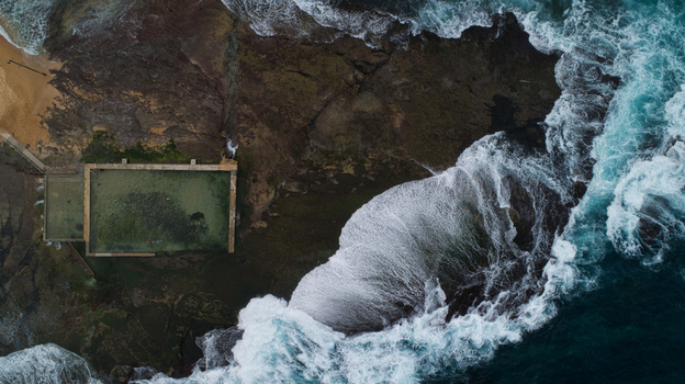 Aerial view of Newport Rock Pool with people swimming as waves crash against the rugged coastline and dark rocks in Sydney New South Wales Australia.
