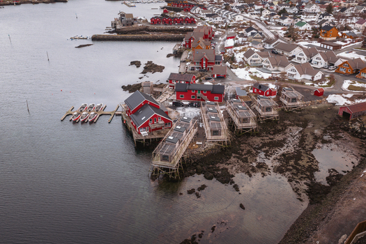 Aerial view of red cabins meet the steely sea under a muted sky contrasting against the town nestled in snow Ballstad Nordland Norway. Aerial view of red cabins meet the steely sea under a muted sky contrasting against the town nestled in snow Ballstad Nordland Norway.