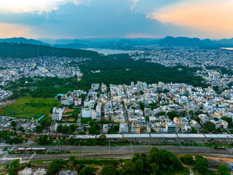 Aerial view of the urban landscape featuring dense buildings lush green spaces distant hills and a railway line in the foreground under a sunset sky in Udaipur Rajasthan India. Aerial view of the urban landscape featuring dense buildings lush green spaces distant hills and a railway line in the foreground under a sunset sky in Udaipur Rajasthan India.
