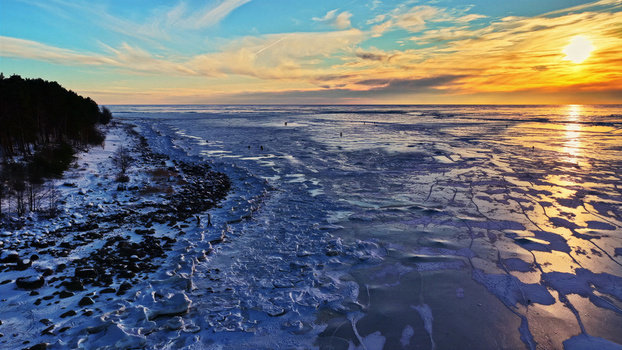 Aerial view of a frozen coastline with ice covered sea dark forest and people walking on the ice during a vibrant golden hour sunset in Latvia. Aerial view of a frozen coastline with ice covered sea dark forest and people walking on the ice during a vibrant golden hour sunset in Latvia.