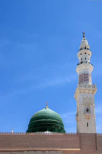 View of the iconic green dome and a tall white minaret of Al Masjid e Nabawi under a clear blue sky in Madinah Al Madinah Province Saudi Arabia.