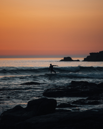 Penzance England United Kingdom 18 July 2021 View of a surfer riding a wave at Gwenver Beach during a vibrant golden hour sunset with dark rocky foreground and calm sea.