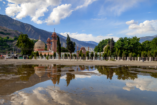 View of the historic Shahi Mosque with its domes and minarets reflected in a large puddle set against a backdrop of mountains and a clear blue sky in Chitral Khyber Pakhtunkhwa Pakistan.