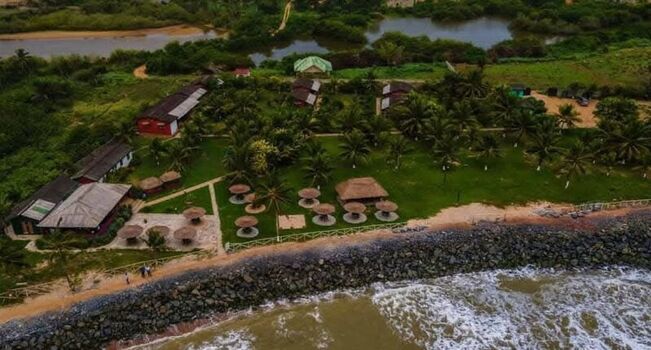 Aerial view of a coastal resort featuring thatched roof huts lush green lawns with palm trees and a rocky shoreline along the Atlantic Ocean in Ghana. Aerial view of a coastal resort featuring thatched roof huts lush green lawns with palm trees and a rocky shoreline along the Atlantic Ocean in Ghana.