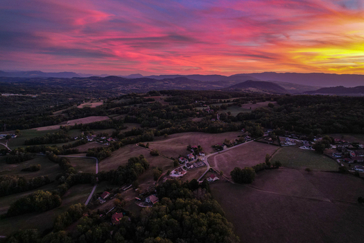 Aerial view of a rural landscape at sunset with vibrant pink and yellow clouds over rolling hills forests and small village houses in France.
