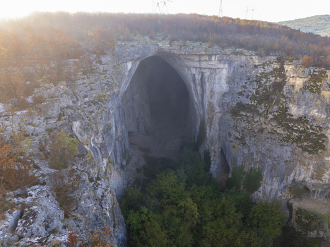 Aerial view of Prohodna Cave featuring its massive limestone arch lush green trees at the entrance and rugged rock formations under a soft morning light in Karlukovo Lovech Bulgaria. Aerial view of Prohodna Cave featuring its massive limestone arch lush green trees at the entrance and rugged rock formations under a soft morning light in Karlukovo Lovech Bulgaria.